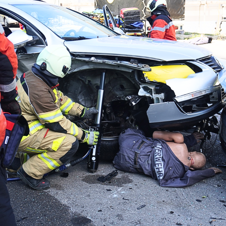 Crash-Test-Dummie liegt unter einem Auto, ein Feuerwehrmann hebt das Auto mit einem LUKAS-Zylinder an